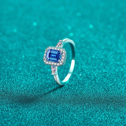 Stunning 1 Carat Moissanite 925 Sterling Silver Ring with a sparkling blue center stone and glittering diamond accents, set against a vibrant teal background.