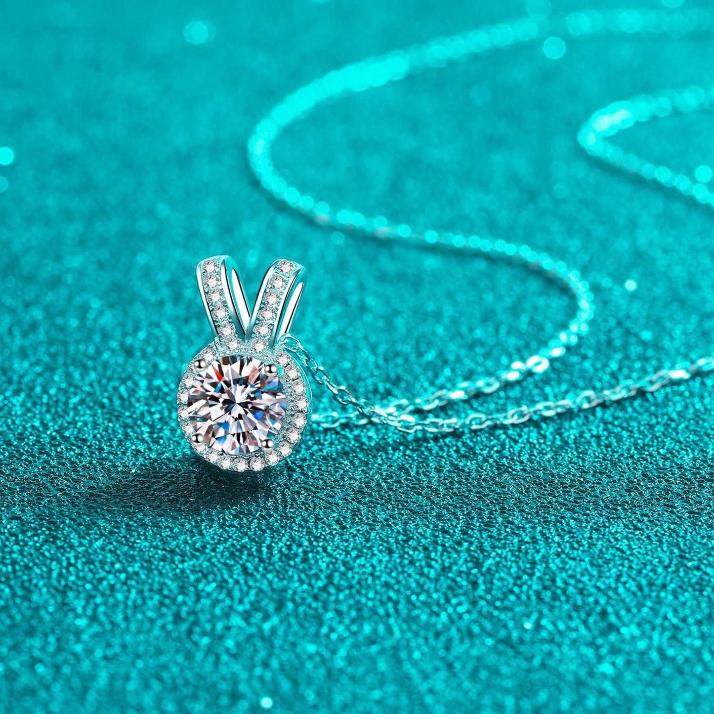 Elegant moissanite pendant necklace with rabbit ear design, sparkling center stone on a metallic chain against a turquoise background.