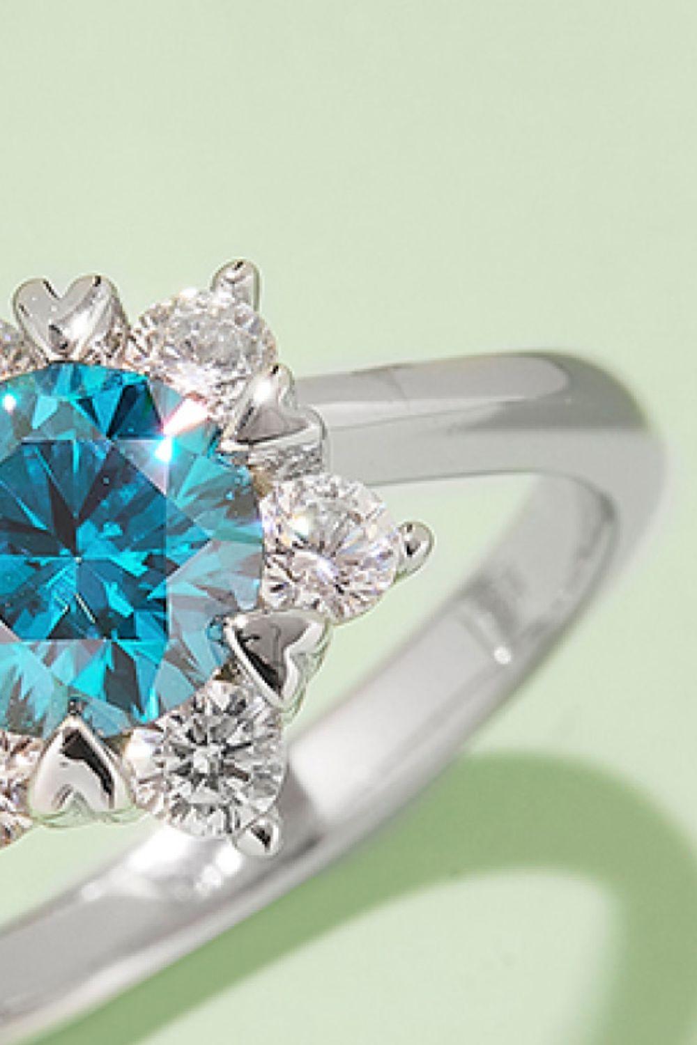 Elegant silver ring with sparkling blue round-cut center stone and surrounding clear stones, displayed against a light green background.