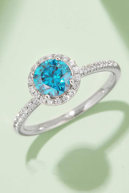 Shimmering silver ring with brilliant blue Moissanite gemstone surrounded by sparkling diamond halo, elegant jewelry piece.