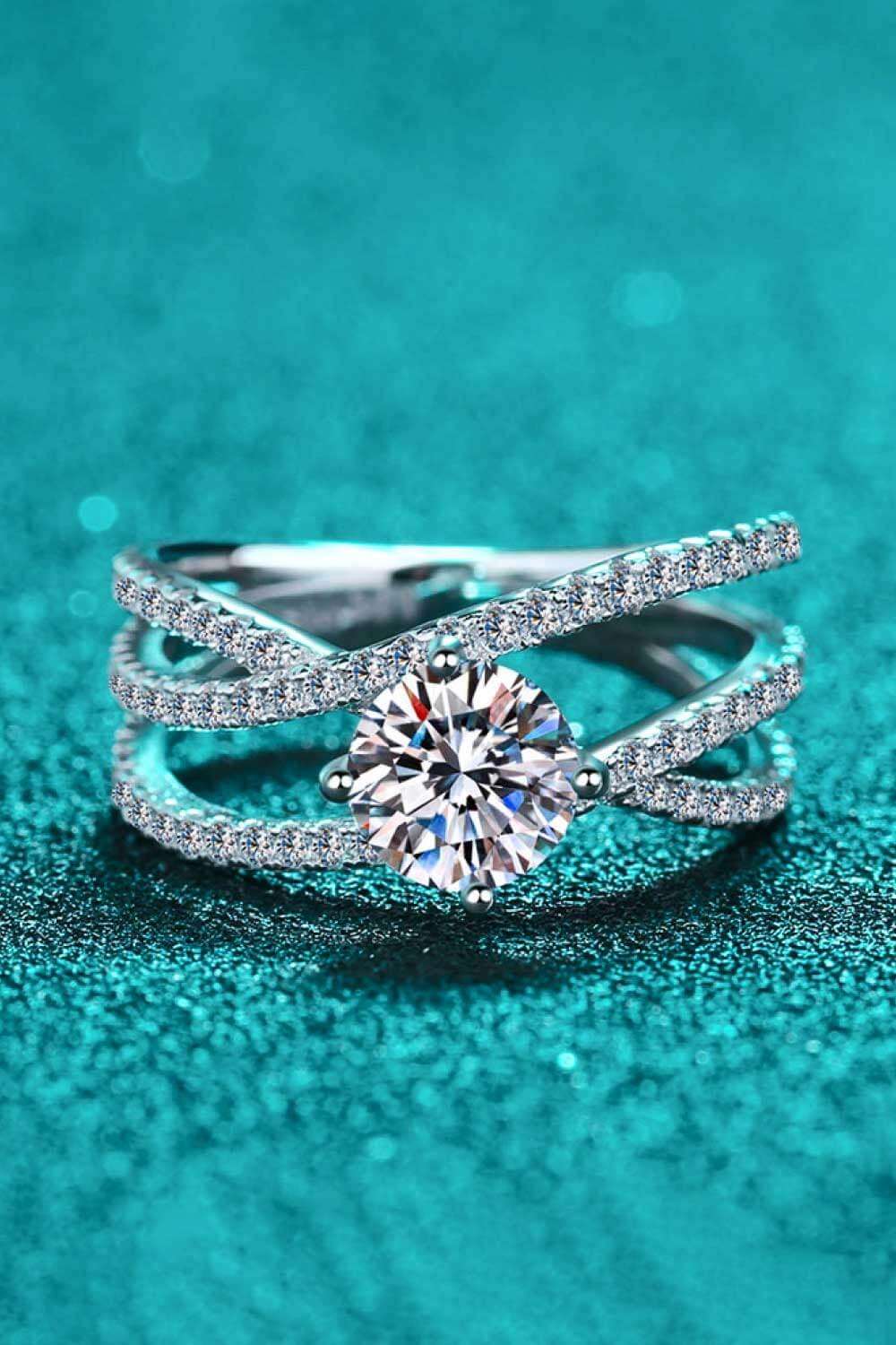 Elegant diamond ring with crisscross design on teal background. The ring features a prominent round-cut gemstone surrounded by a sparkling diamond-encrusted band.