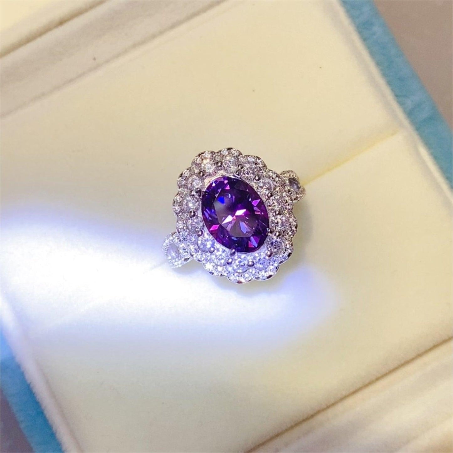 Elegant 2 Carat Moissanite 925 Sterling Silver Ring with a vibrant purple oval gemstone surrounded by a glittering crystal halo, displayed on a soft blue velvet surface.