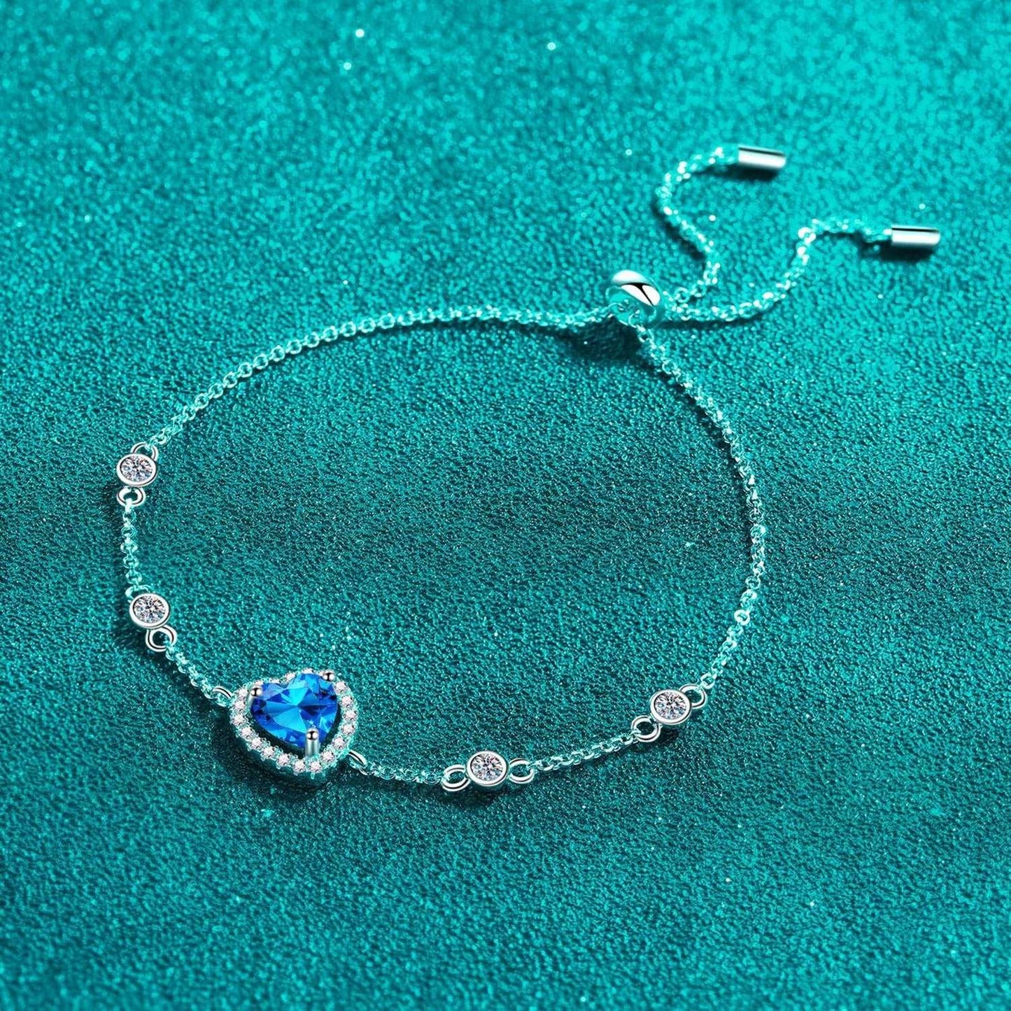 Elegant sterling silver bracelet with heart-shaped blue moissanite center stone and sparkling crystal accents against a vibrant teal background.