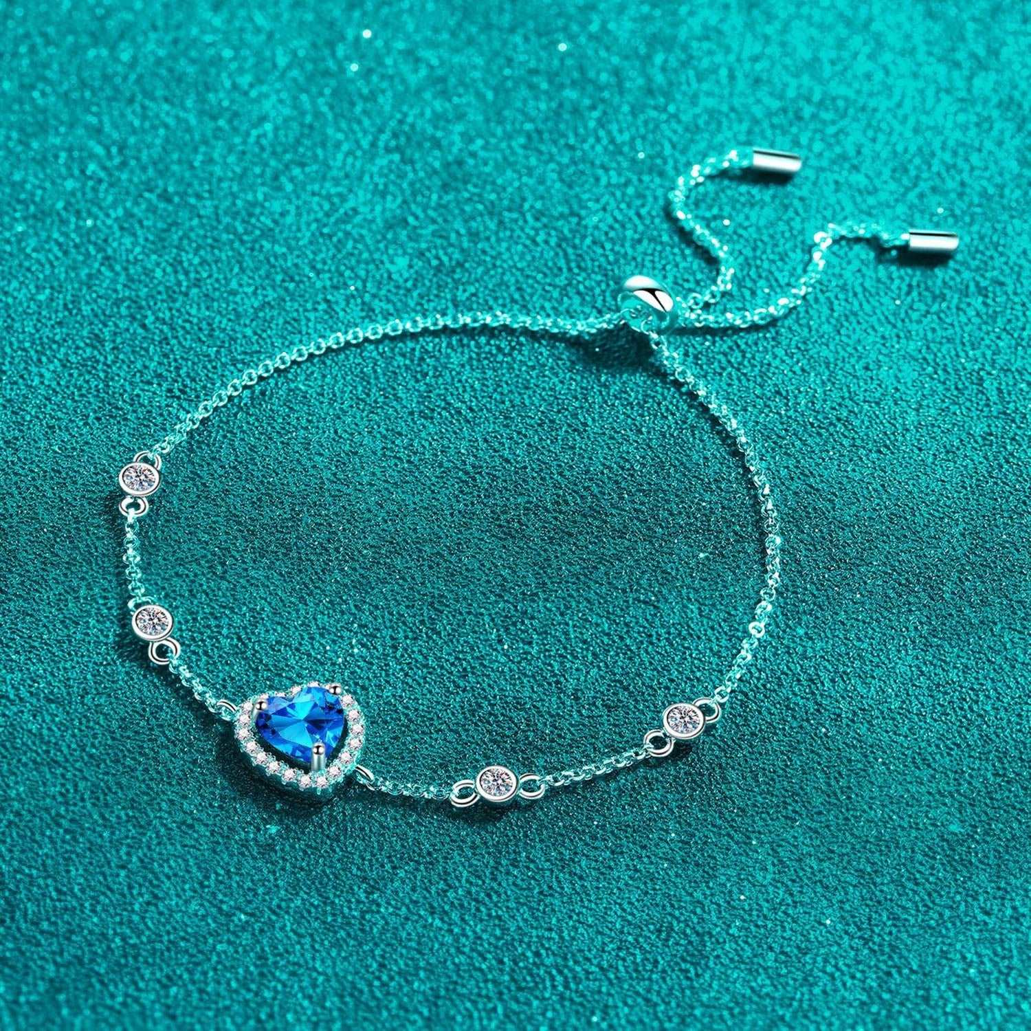 Elegant sterling silver bracelet with heart-shaped blue moissanite center stone and sparkling crystal accents against a vibrant teal background.