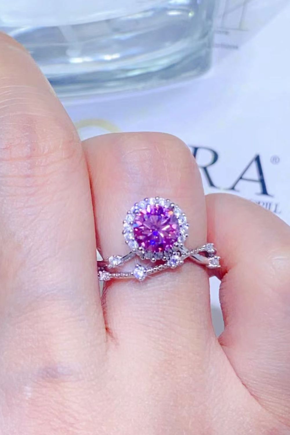 Elegant purple moissanite gem ring with sparkling silver band, resting on a hand against a blurred background.