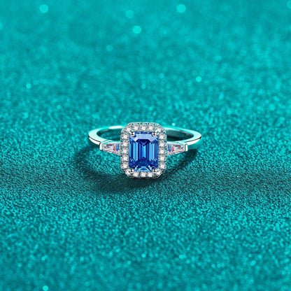 Elegant 1 Carat Moissanite 925 Sterling Silver Ring with Sparkling Emerald-Cut Blue Stone Surrounded by Diamond Halo on Teal Glitter Background