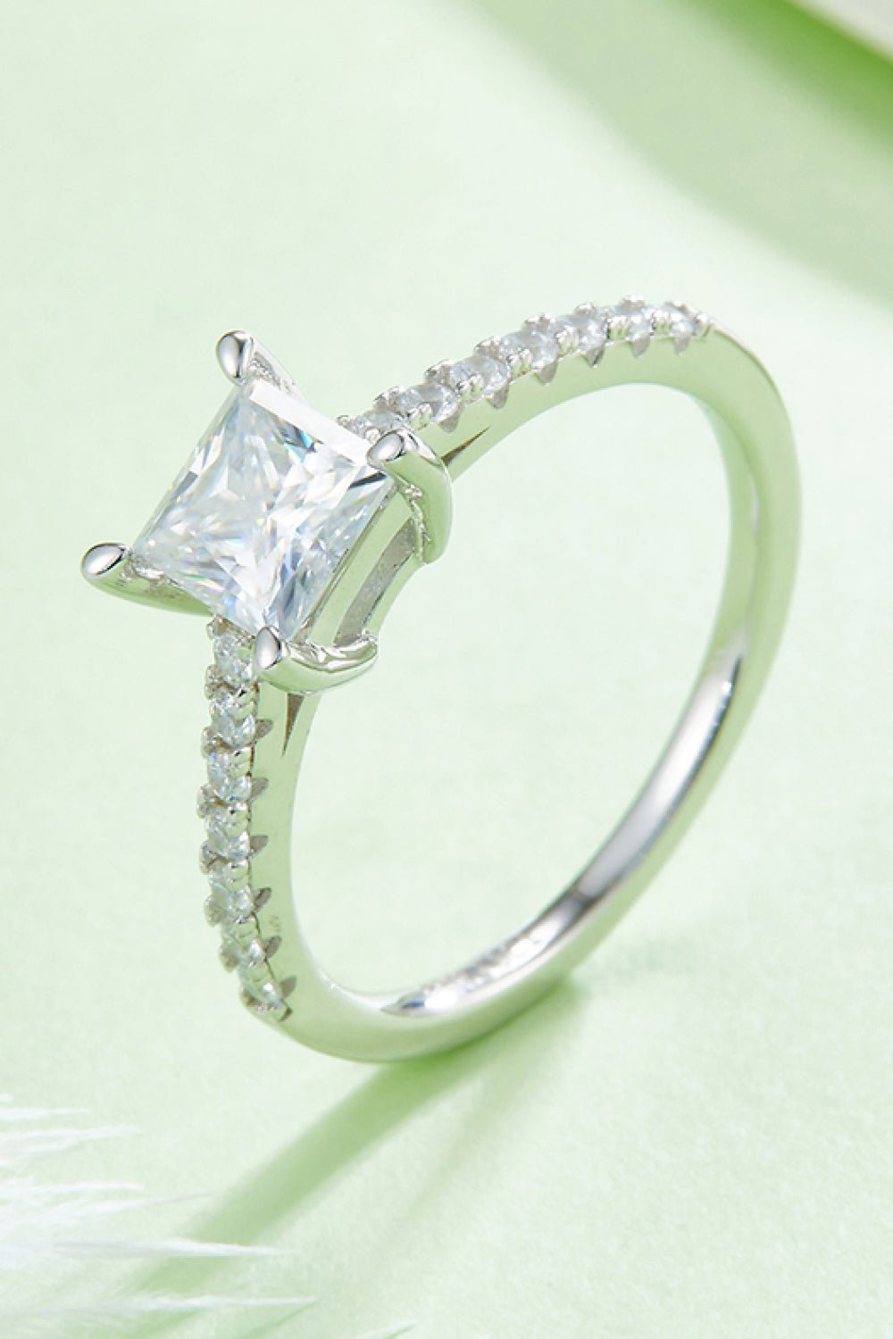 A princess-cut moissanite center stone in a sterling silver ring with sparkling side stones, set against a soft green background.