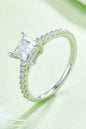 A princess-cut moissanite center stone in a sterling silver ring with sparkling side stones, set against a soft green background.