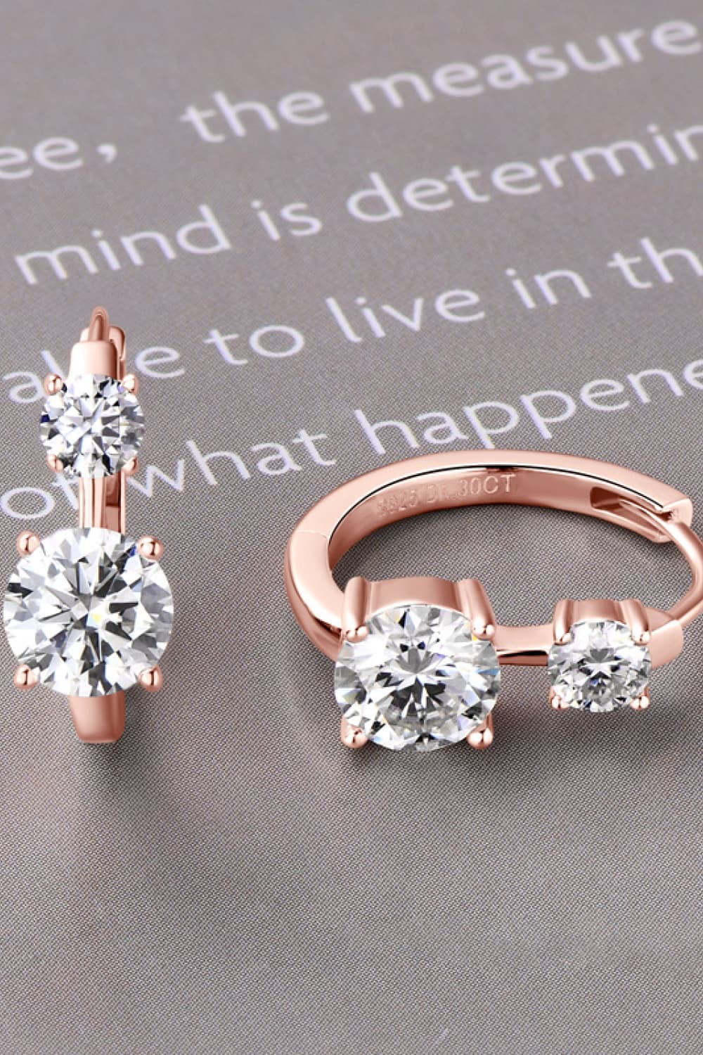 Elegant rose gold diamond earrings and ring displayed on a gray surface with a motivational quote in the background.