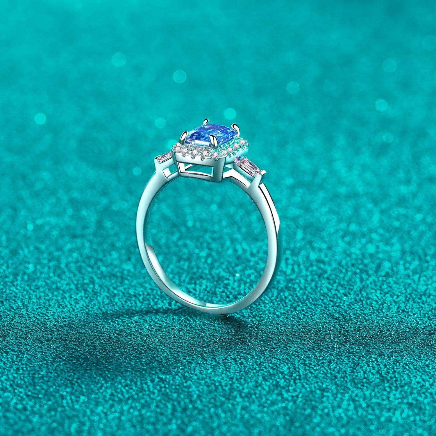 Elegant sterling silver ring with sparkling moissanite center stone surrounded by smaller accent stones, set against a vibrant teal background.