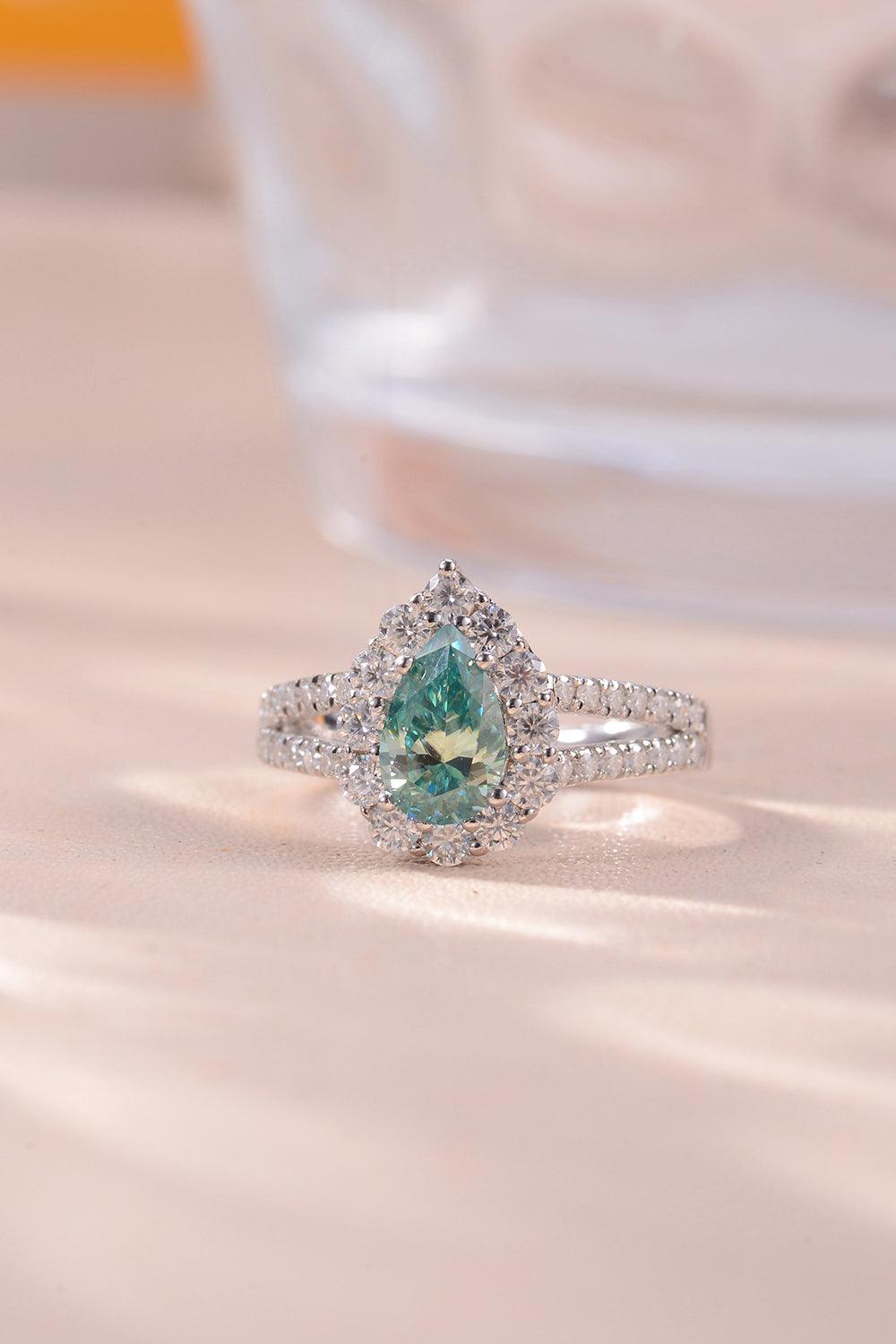 Elegant pear-shaped emerald ring with diamond halo and split shank, showcasing a stunning center stone set in a silver-toned metal band on a soft, beige background.